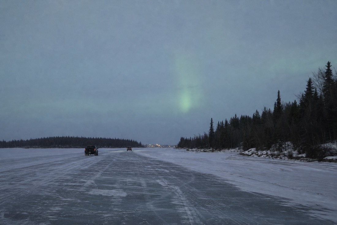Two vehicles keeping a long spacing on the ice road under flat grey light with the Dettah shoreline ahead