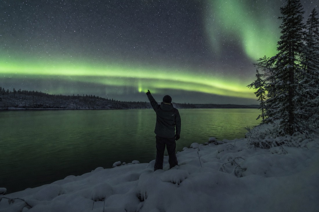 Guide pointing north from a dark lakeshore as a green auroral arc stretches across the whole horizon