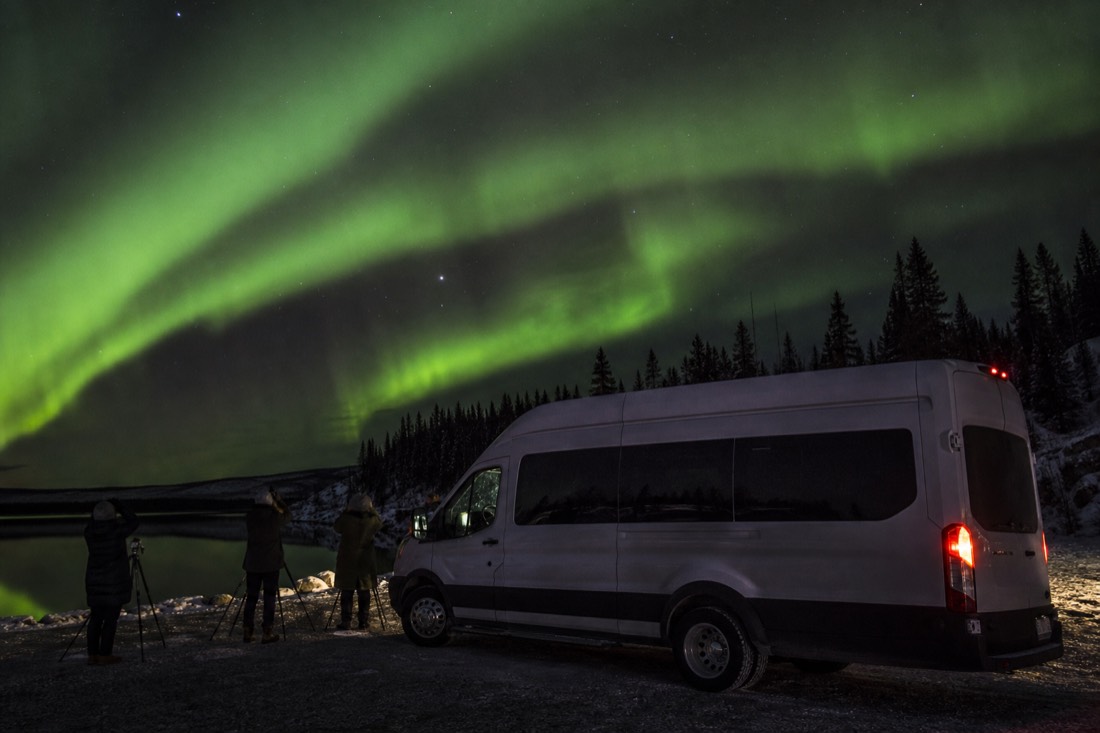 Heated tour van parked at a dark lakeside pullout with guests photographing green aurora overhead
