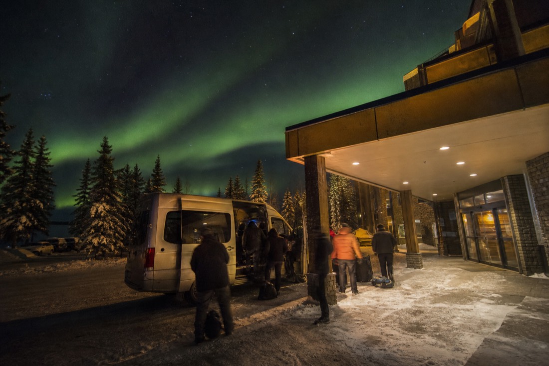 Travellers loading camera bags into a tour van outside a lit Yellowknife hotel entrance on a snowy night