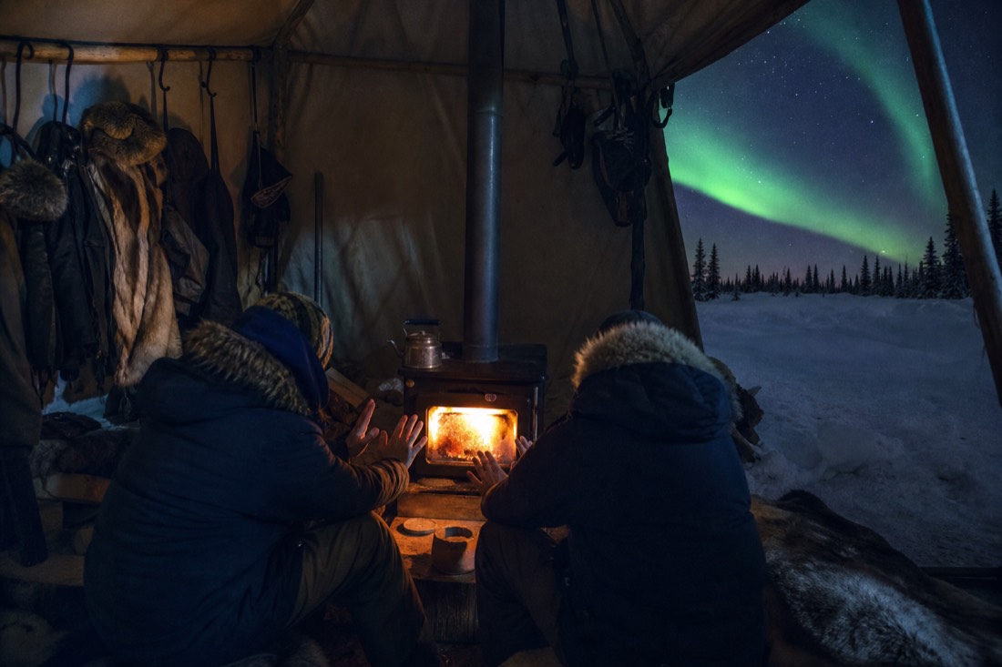 Guests warming hands around a wood stove inside a canvas shelter with winter gear hanging nearby