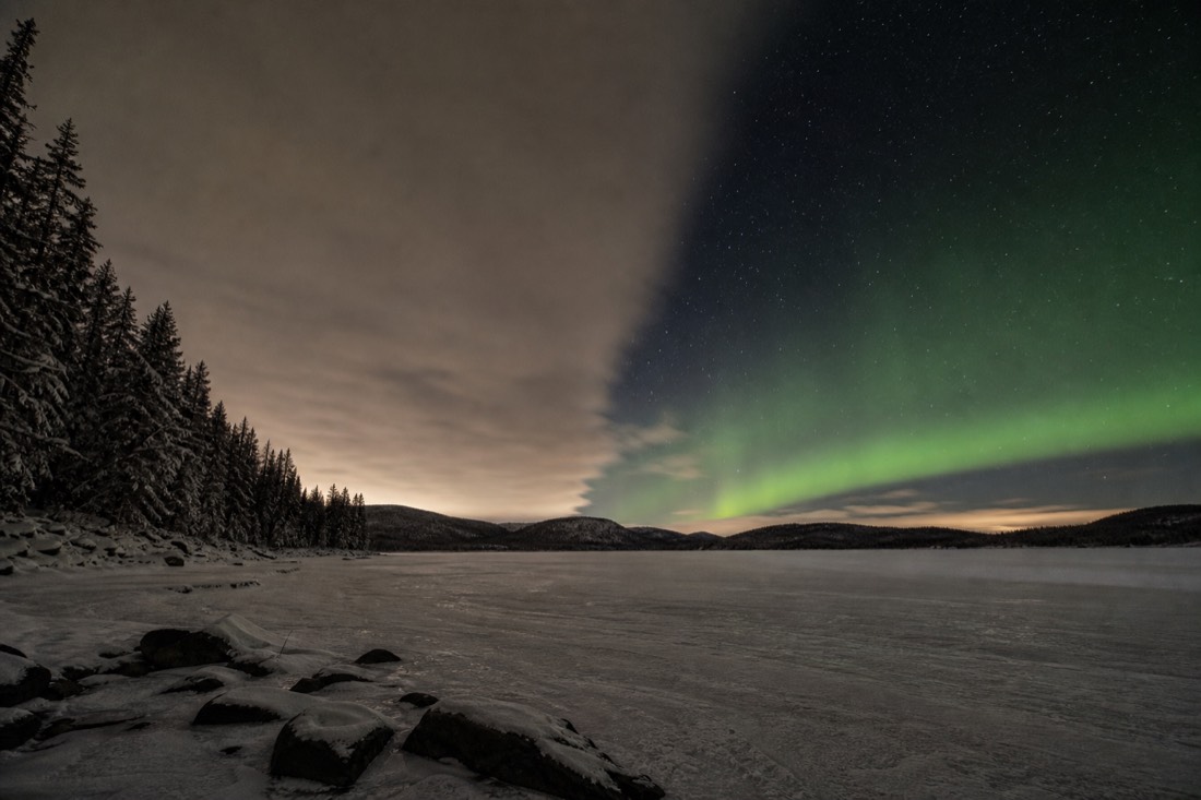 Broken cloud over a frozen lake with green aurora showing through a clear gap on one side of the sky