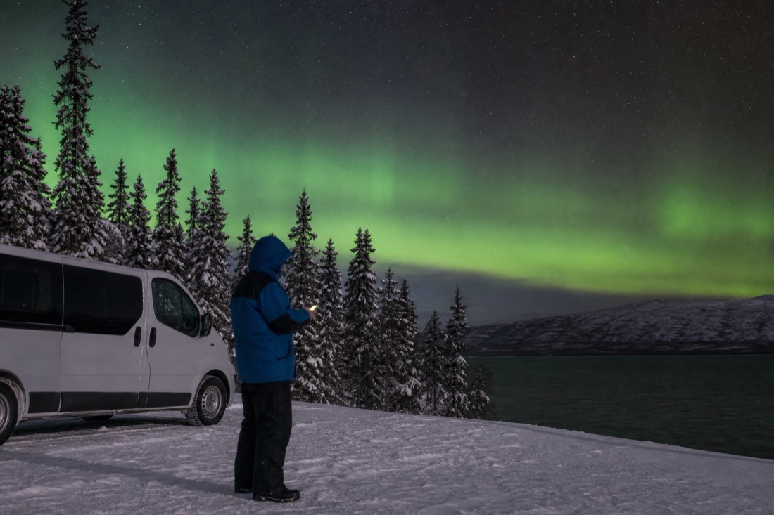 Tour guide in heavy arctic clothing checking a phone beside a parked van on a snowy lakeshore at night
