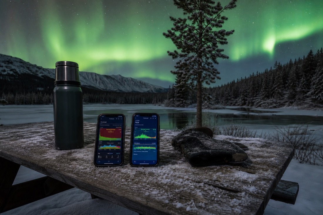 Two phones on a snow-covered picnic table showing different aurora app screens beside a thermos and mittens