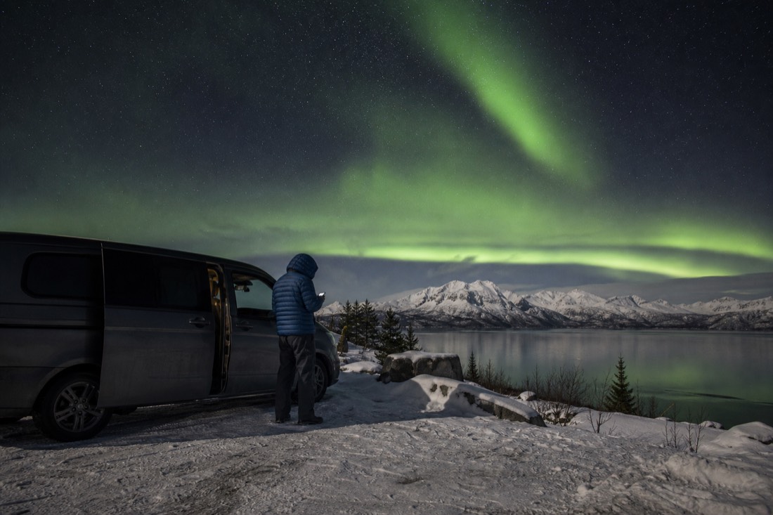 Guide checking a phone forecast beside a parked van on a snowy lakeside pullout under a starry sky