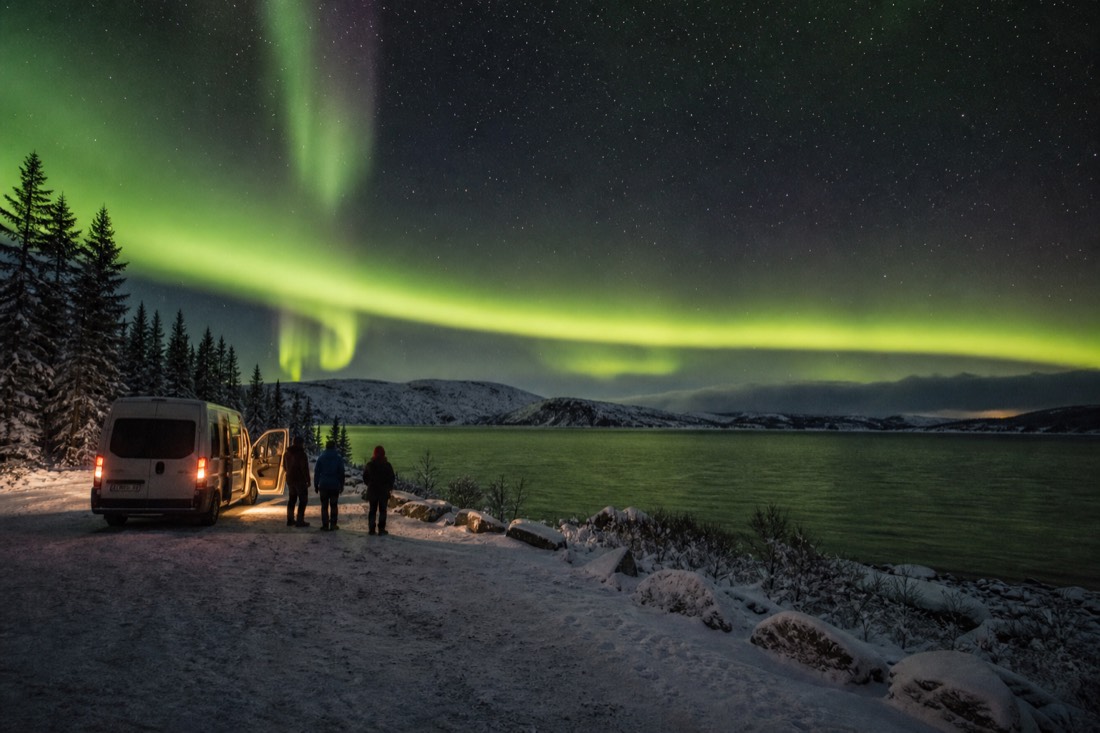 Small group standing beside a heated van on a lakeside viewpoint watching green aurora overhead