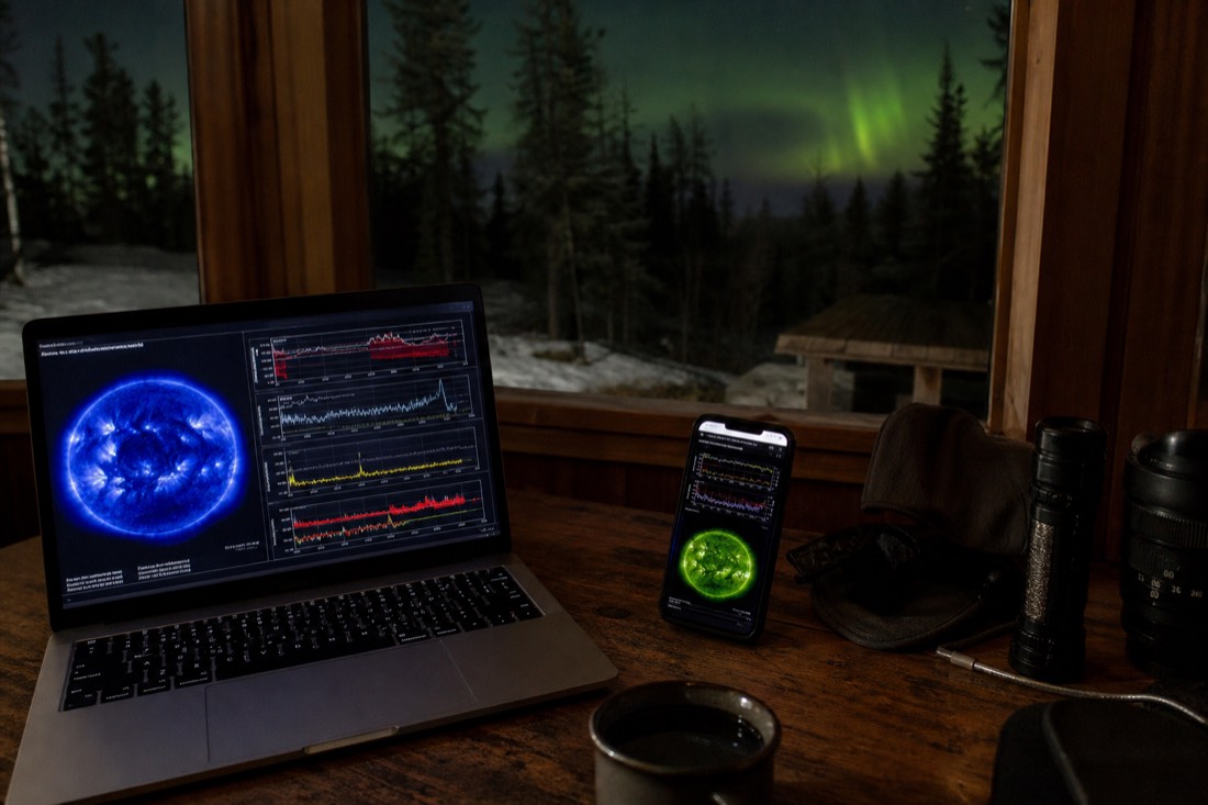 Laptop and phone showing space weather charts on a cabin table with a dark window and faint green sky behind