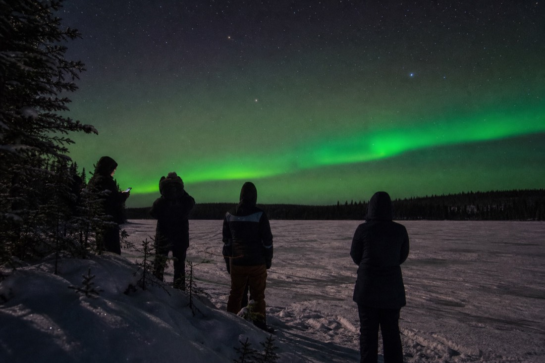 Group of watchers on a frozen lakeside at night, one glancing at a dimmed phone screen while green aurora rises behind