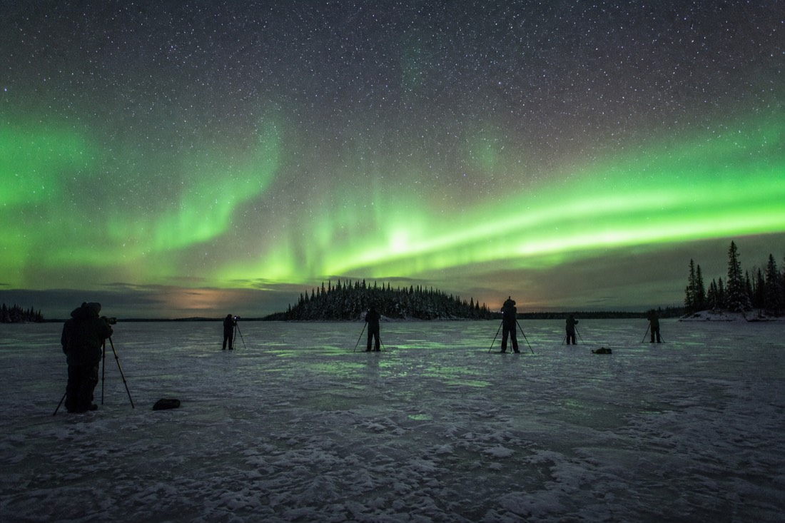 Aurora watchers with tripods spread across open lake ice under a wide star-filled northern sky
