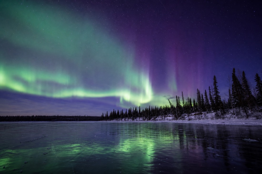 Green and violet aurora reflected on smooth lake ice with a low horizon of dark spruce forest