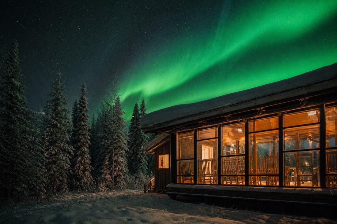 Warmly lit heated viewing lodge windows glowing under a green aurora sky in northern forest
