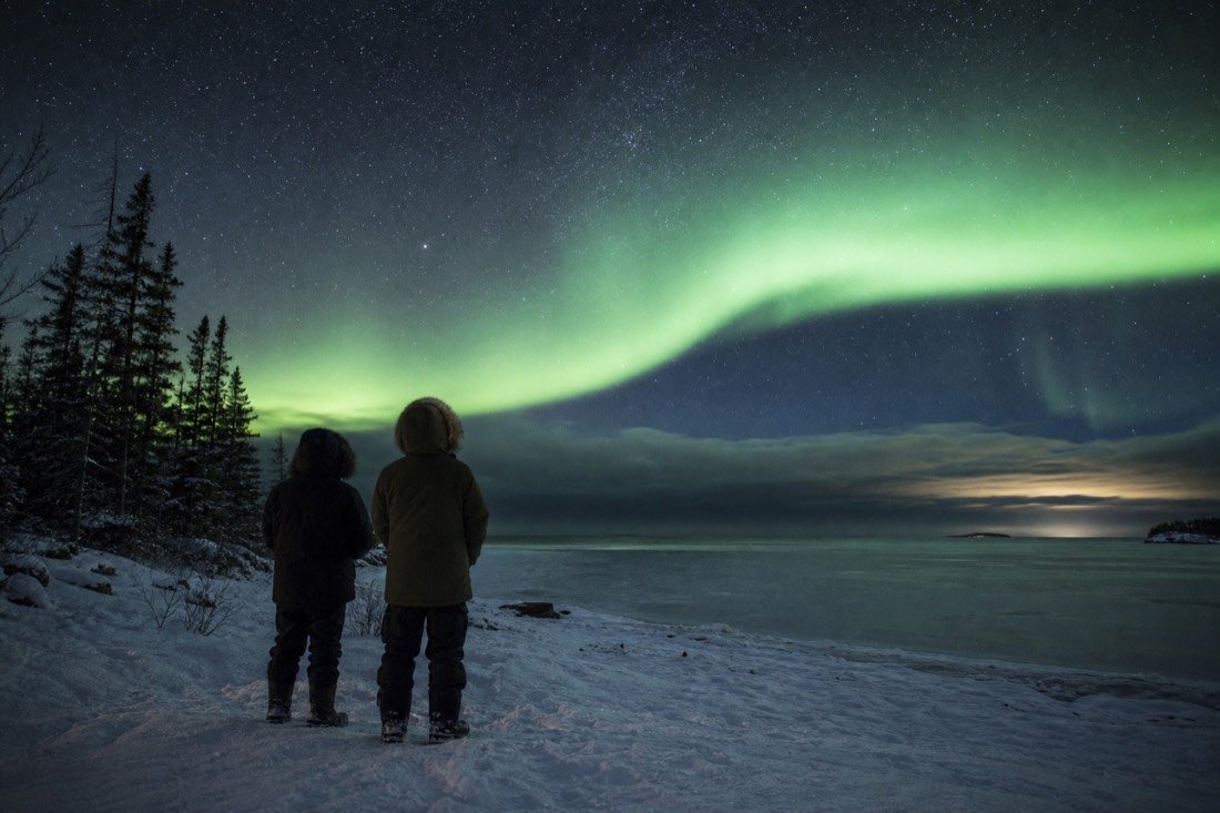 Two travellers in heavy parkas watching a pale green aurora arc from a snow-covered northern shoreline