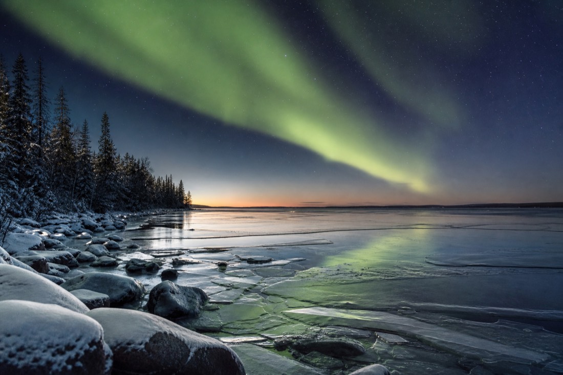 Half-frozen northern lake at dusk during freeze-up with thin ice sheets forming near a rocky shoreline