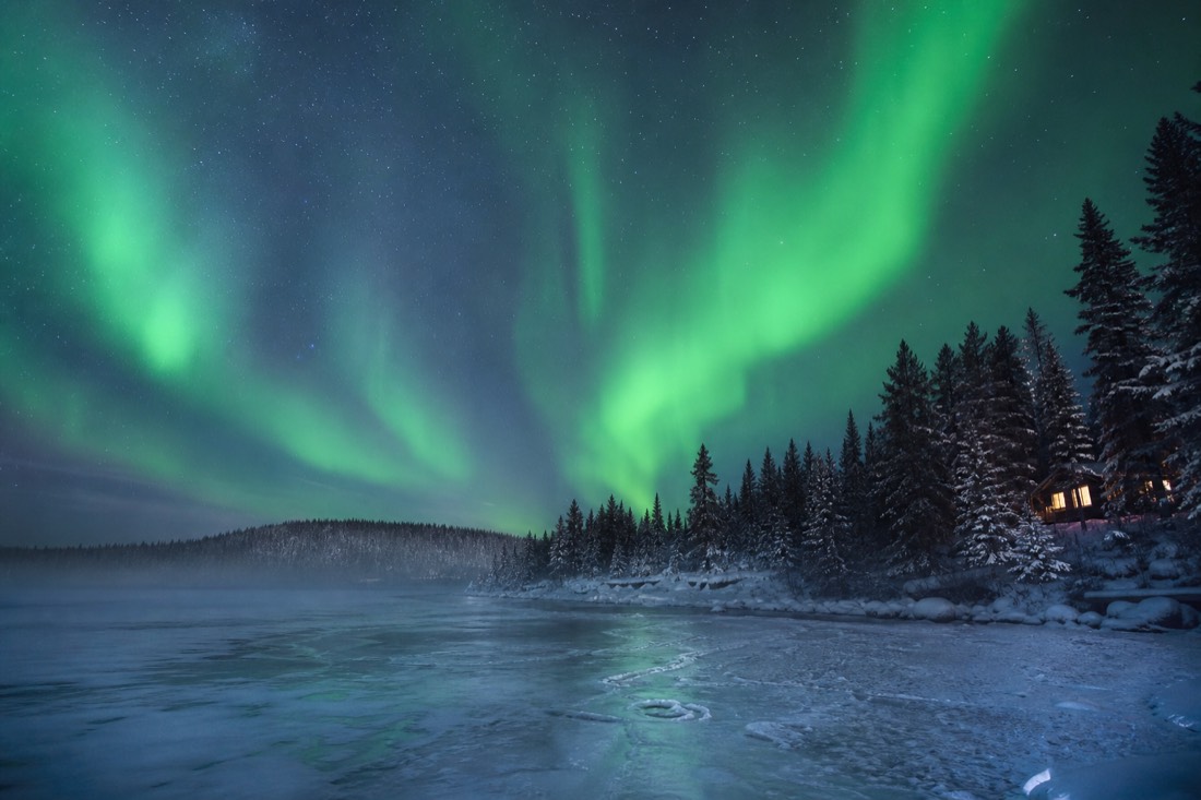 Aurora curtains over a frozen lake at the edge of a spruce forest with a lit cabin window in the distance