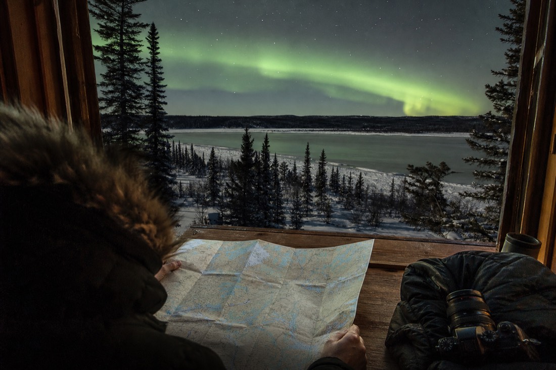 Traveler studying a paper map of northern Canada beside a parka and camera on a wooden cabin table