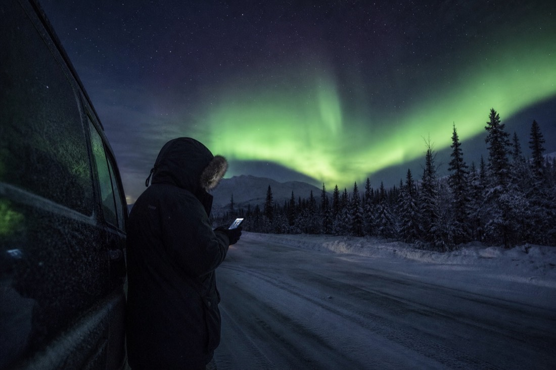 Guide in a heavy parka checking a weather forecast on a phone beside a parked van on a dark snowy road