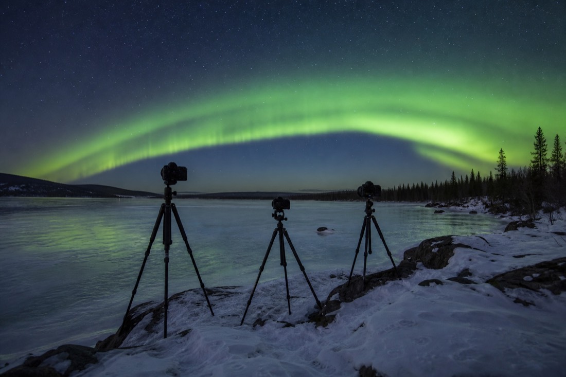 Three tripods silhouetted on a frozen lake shore beneath a bright arcing aurora band