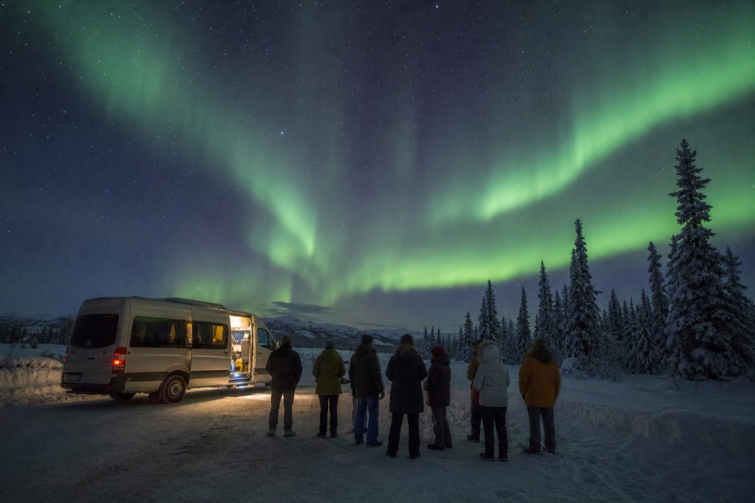 Small tour group waiting under a starry sky beside a heated van on a snowy northern road