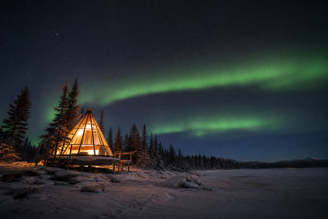 Glass-roofed tipi glowing warm on a frozen lake beneath a green aurora band