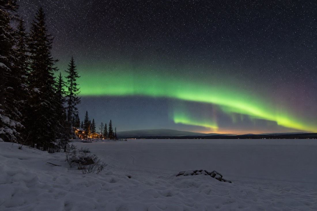 Long horizon-to-horizon aurora arc above a snow-covered lake with distant cabin lights