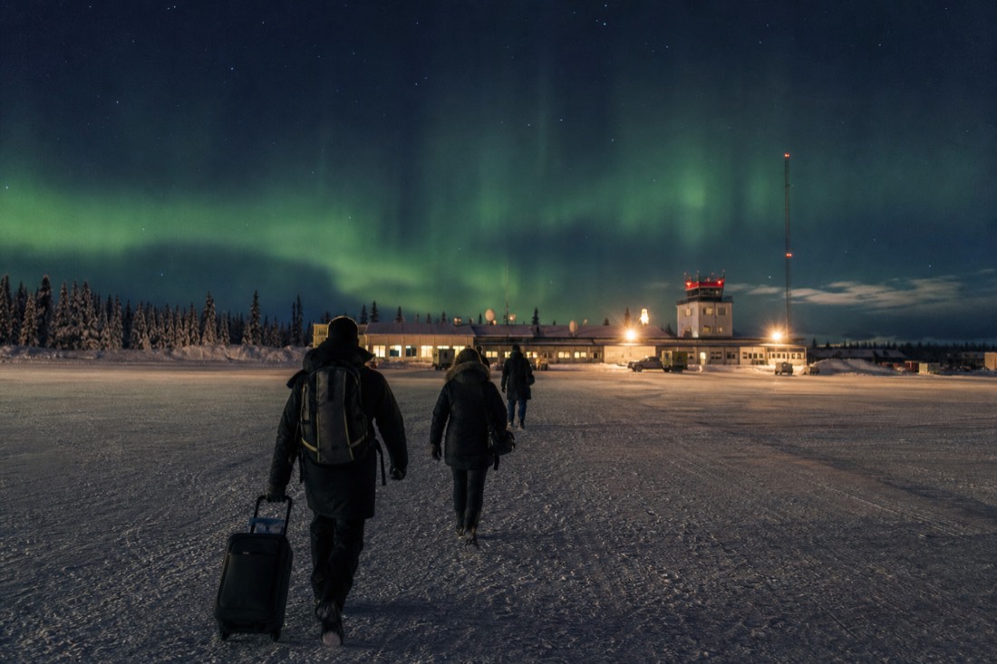 Travelers walking across a snowy airport apron toward a small terminal building at dusk in northern Canada