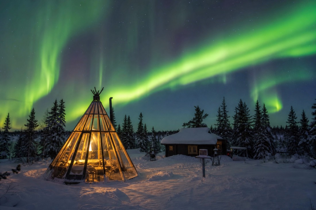 Glass tipi glowing warmly under a bright aurora display at a wilderness cabin camp near Yellowknife