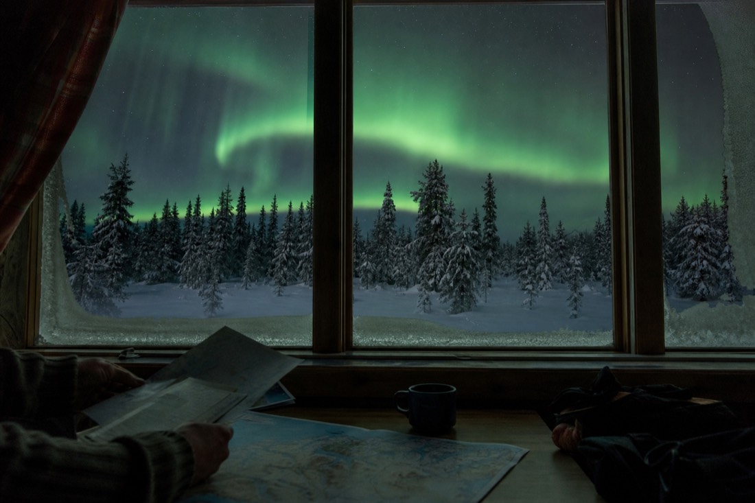Traveler comparing trip notes by a frosted window with green aurora glowing over the snow outside