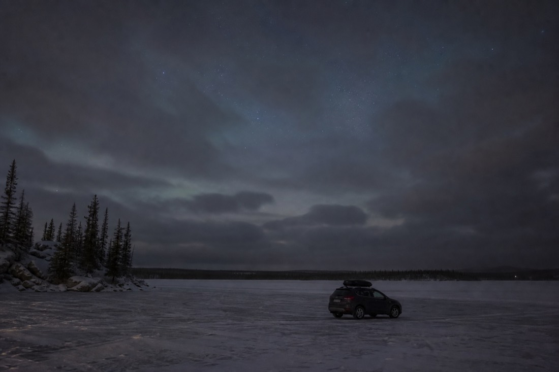 Empty cloudy night sky over a dark frozen lake with a single parked vehicle and no aurora visible