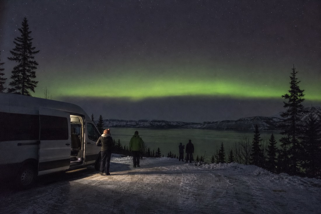 Heated tour van parked at a dark lakeside viewpoint with guests stepping out under a faint aurora arc