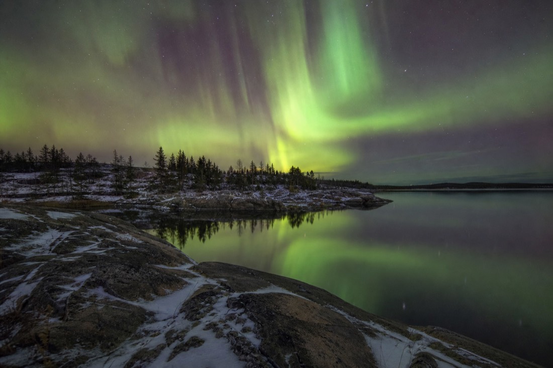 Autumn aurora reflected in the open water of a lake near Yellowknife with bare rock in the foreground