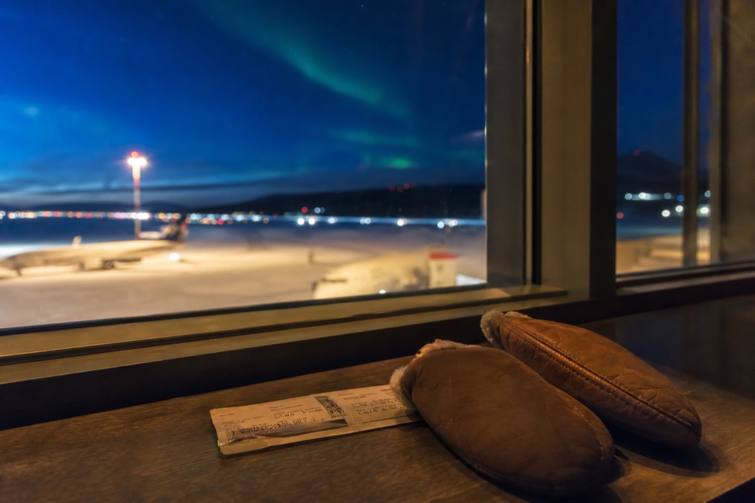 Boarding pass and warm mitts on a table beside a window showing a snowy northern runway at dusk