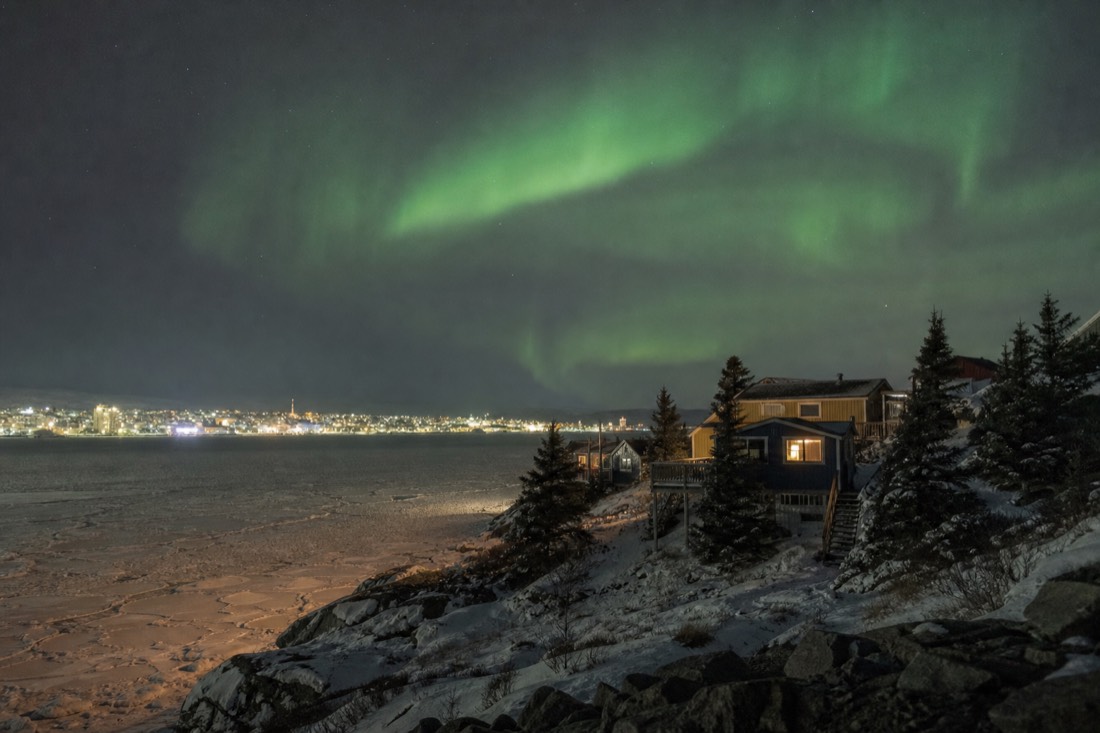 Small northern community of low wooden houses on a rocky shore across a frozen bay from a distant town skyline