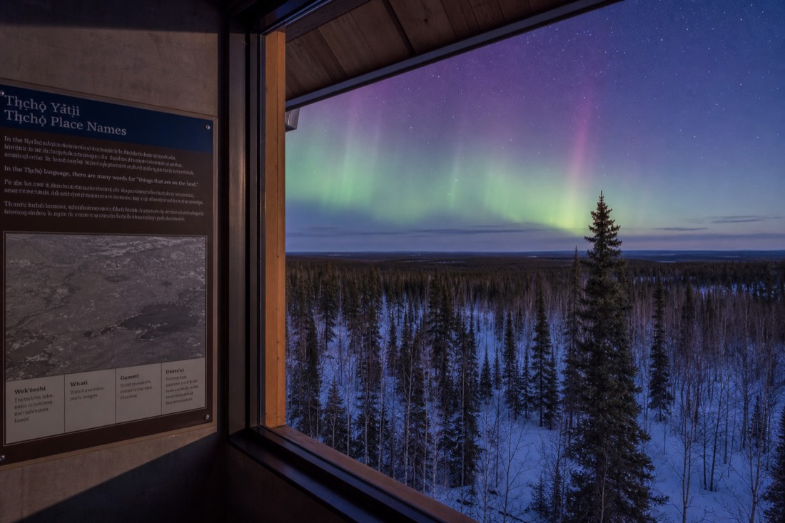 Multilingual interpretive panel of northern place names mounted beside a window looking onto boreal forest