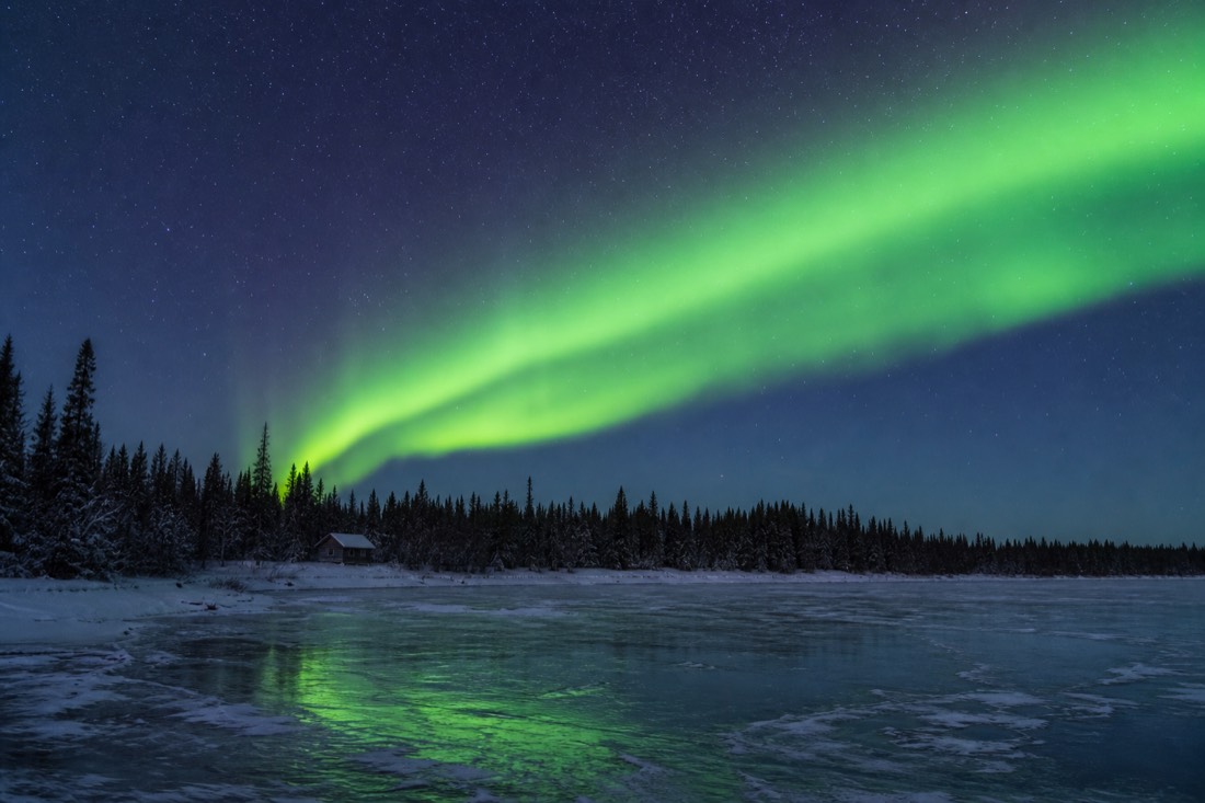 Green aurora arc over a frozen lake and black spruce treeline with a small unlit cabin in the distance