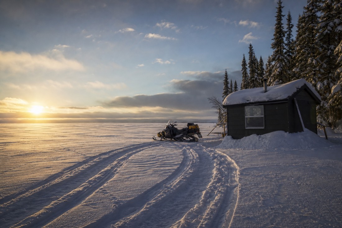 Heated fishing hut and snowmobile track on a wide frozen lake under low winter sun