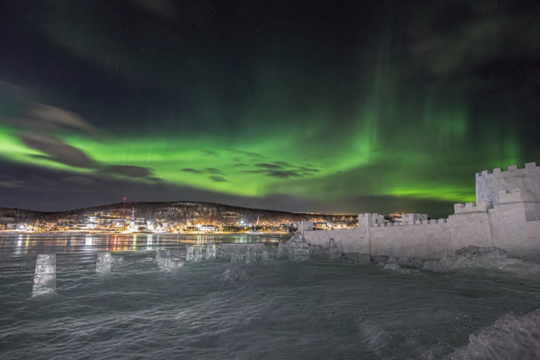 Snow castle walls and carved ice blocks on a frozen bay with a town shoreline behind
