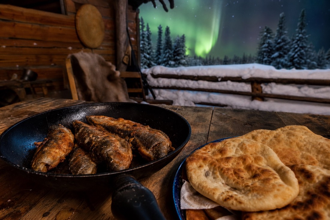 Pan of fresh bannock and fried lake fish on a plank table inside a warm wooden cabin