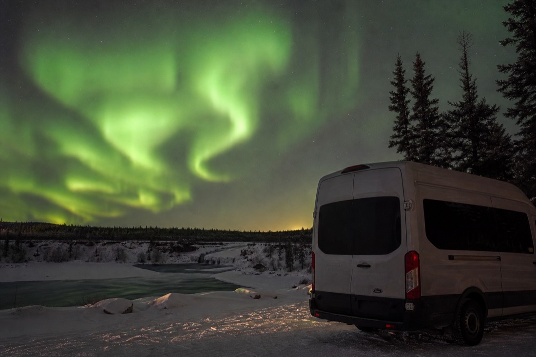 Heated tour van parked at a dark lakeside viewpoint under a bright green aurora near Yellowknife