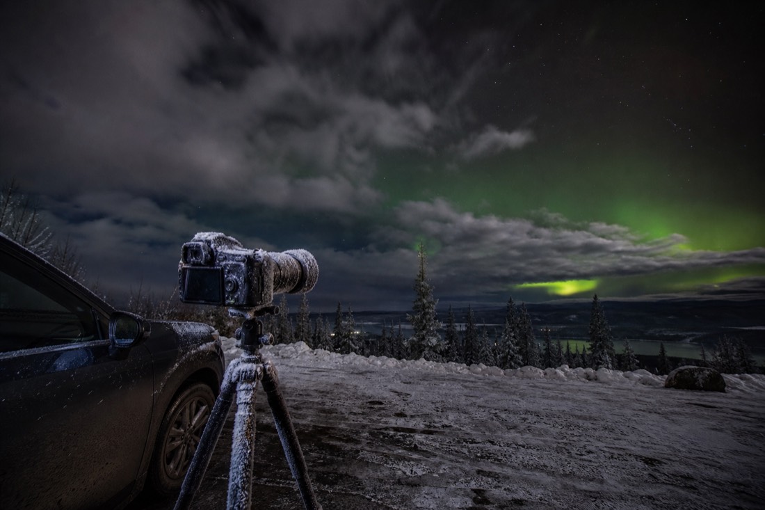 Frost covered camera on a tripod beside a parked car at a dark roadside pullout with heavy cloud over most of the sky