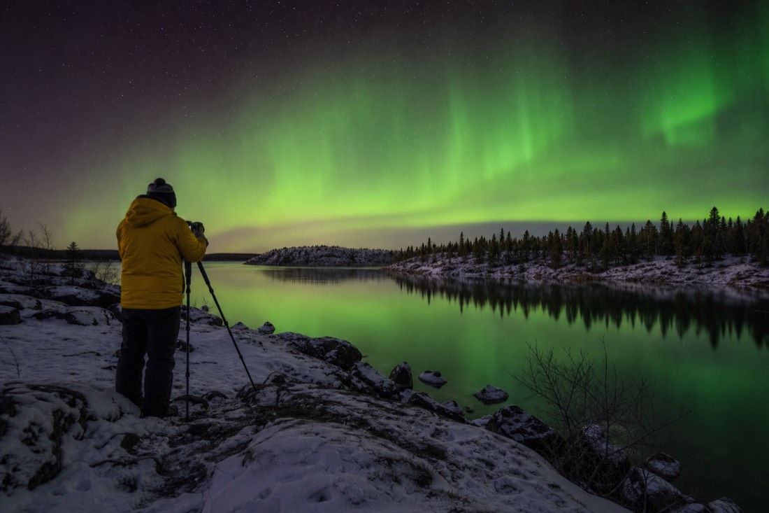 Traveller in autumn jacket photographing green aurora reflected in open lake water on a rocky shoreline near Yellowknife