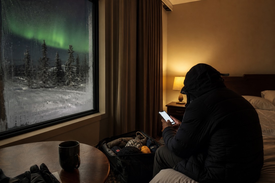 A traveler checking flight dates on a phone beside a packed bag in a warm Yellowknife hotel room with aurora showing through the frosted window