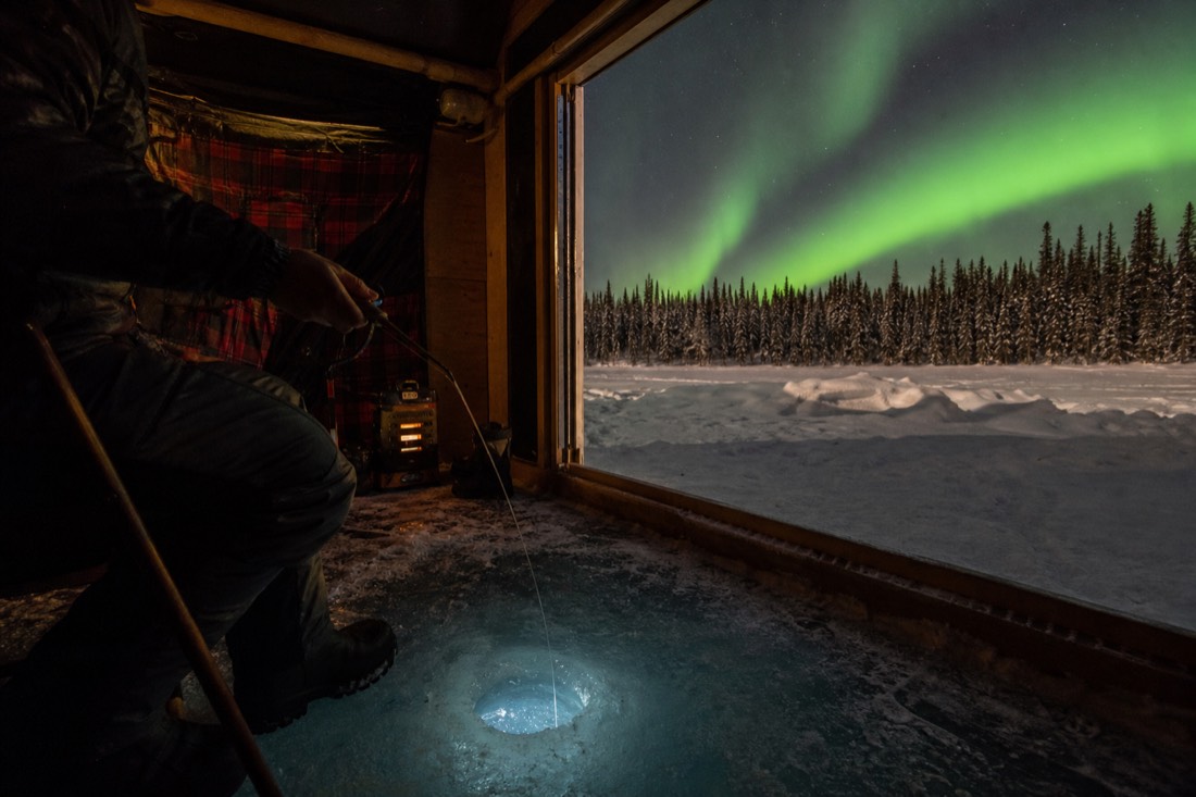 Angler jigging a line through a drilled hole in thick blue lake ice inside a warm fishing hut