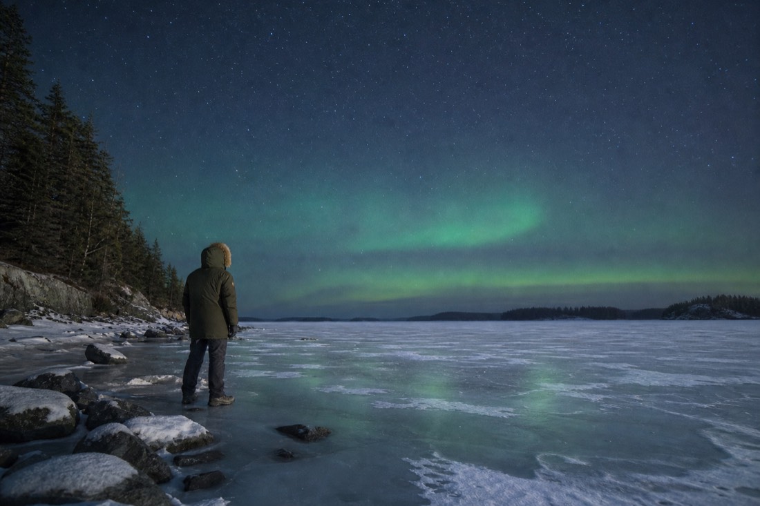 Traveller in a heavy parka standing on a frozen lake looking up at a faint green aurora band