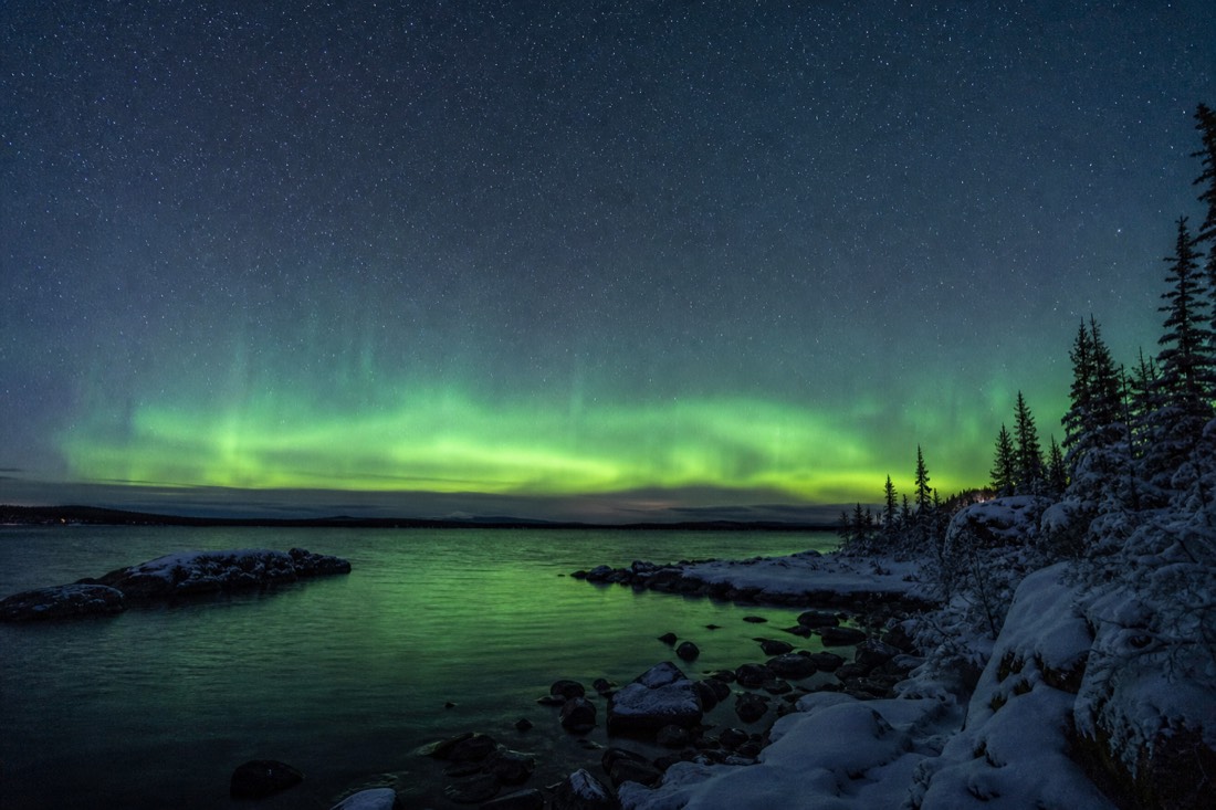 Late August night sky over open water with a pale green aurora glow low above the treeline
