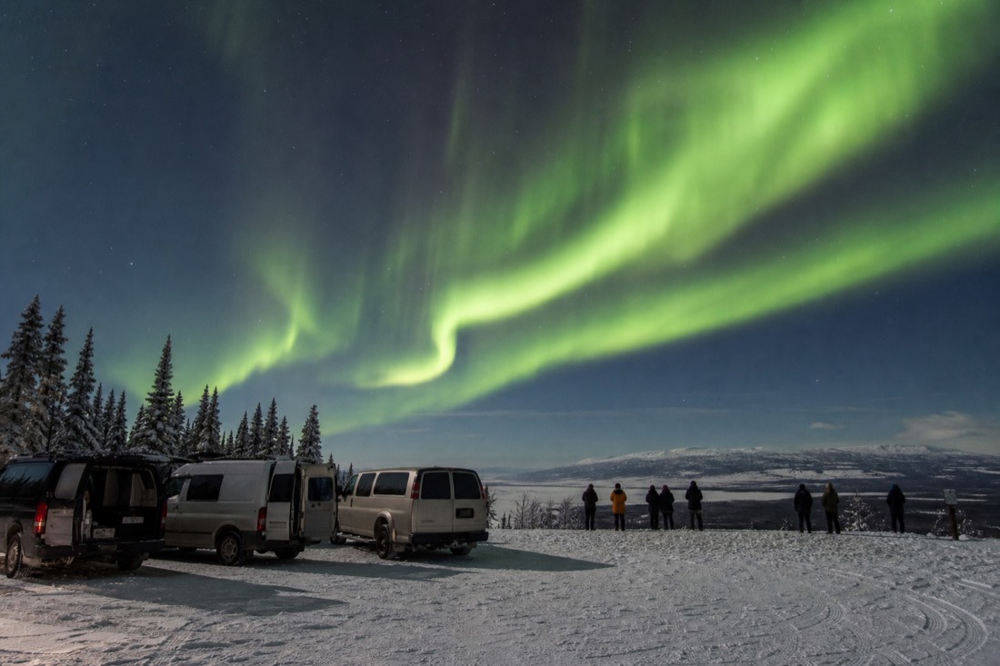 Three parked vans on a snowy lookout with guests watching a bright aurora display overhead