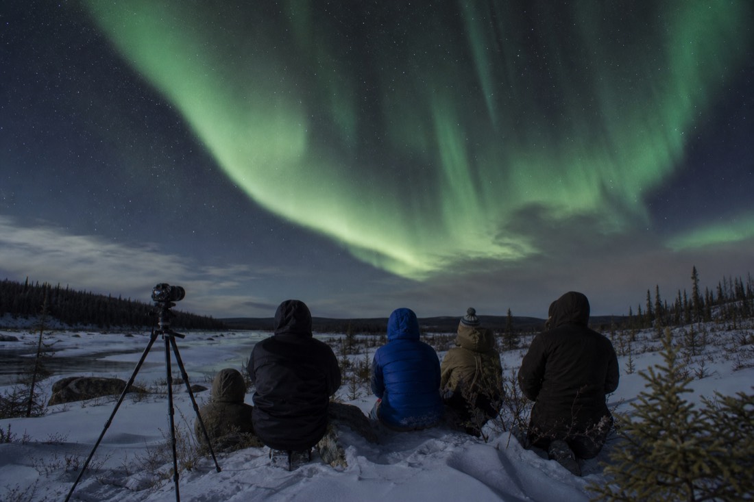 Group of people watching a pale green aurora curtain shift overhead while one camera sits on a tripod