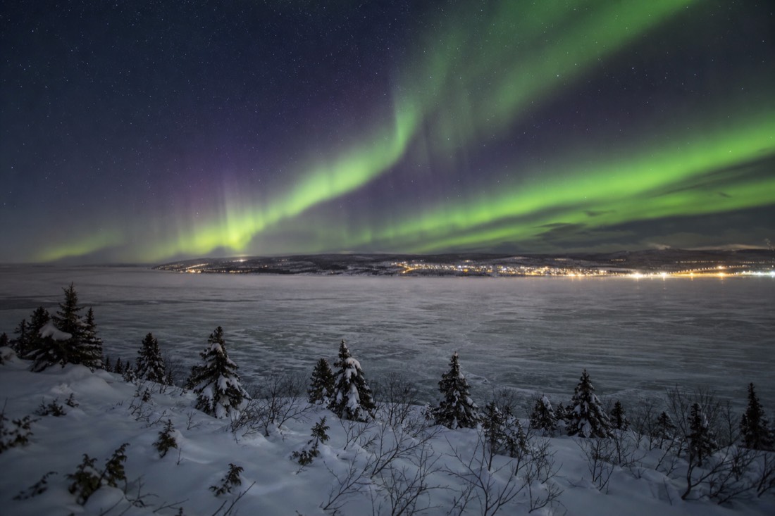 Aurora over a small northern town's lights seen from a snowy ridge above a frozen bay