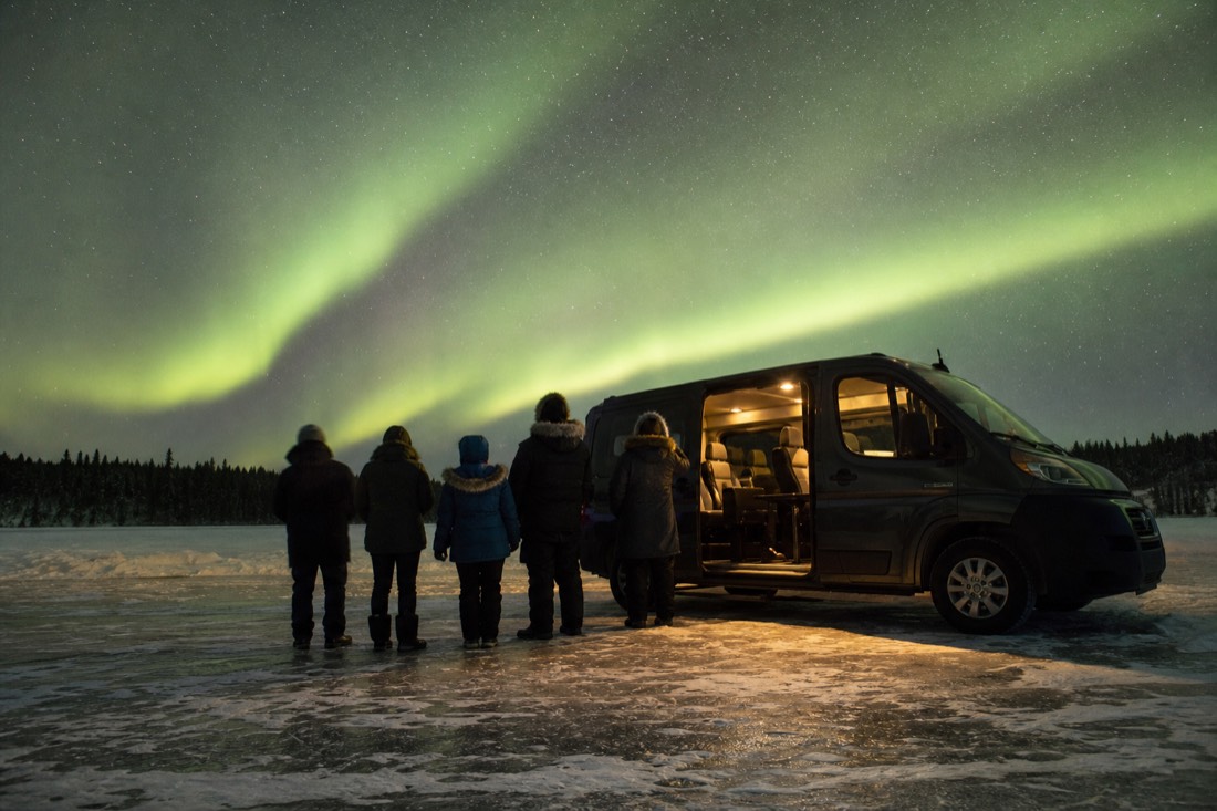 Guests in rental parkas standing beside a heated van on a frozen lake at night waiting for the aurora