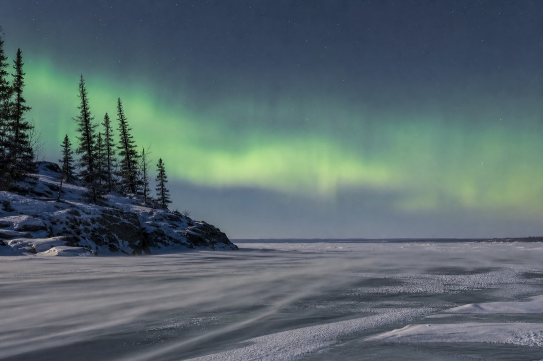 Wind-blown snow drifting across the frozen surface of Yellowknife Bay under a pale winter sun