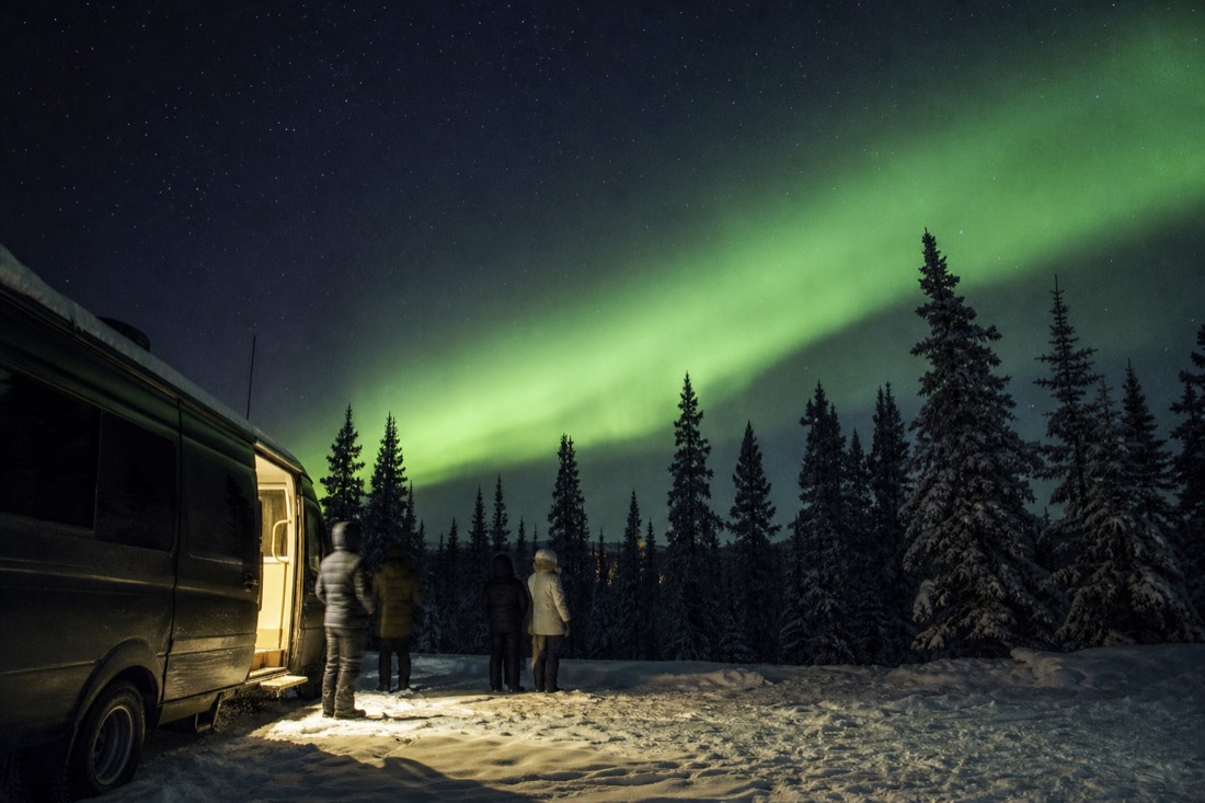 Small group beside a heated tour van watching a green aurora band over dark spruce near Yellowknife